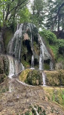 The video shows a waterfall cascading over moss-covered rocks in a wooded area. The water flows in multiple streams down the textured rock face. The surrounding environment is lush with green foliage and trees. The lighting suggests it is daytime, possibly overcast. There are no people or man-made objects visible in the frame.