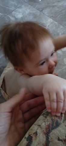 A close-up, top-down video shows a baby lying on its back on a pale, patterned floor. A hand is holding the baby's arm. The baby is wearing a tan onesie and has light brown hair. The baby is looking towards a beige and green patterned sofa cushion. The baby's mouth is partially open, and it appears to be gumming or chewing on the cushion. The video is shaky, suggesting it was filmed with a handheld device.