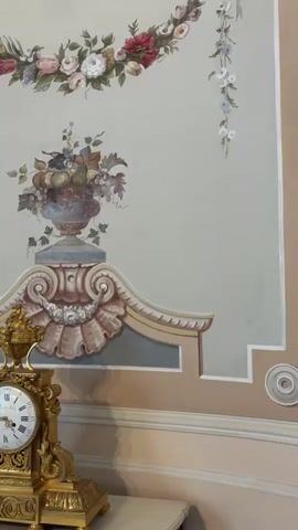 The video shows a wide shot of a room with ornate decorations. The walls are painted in pastel colors with floral motifs and architectural details. In the foreground, there is a gilded antique clock on a table. The camera pans to the right, revealing a window with heavy curtains and intricate moldings. Further to the right, a woman in a brown dress walks away from the camera, holding a phone and a purse. The room appears to be a richly decorated interior, possibly a historic building or a museum.