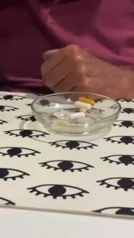 An elderly man with white hair takes pills from a small glass bowl and drinks from a glass. He is wearing a maroon polo shirt. The bowl is filled with various pills and sits on a white table with black eye patterns. Another man in a green shirt enters the frame to place a can on the table. The room's background features a television, a chair, and a speaker.
