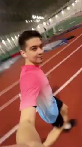 The video shows a young man jogging on an indoor track. He is wearing a pink and blue t-shirt, black shorts, and running shoes. He also has a watch on his left wrist and earbuds in his ears. The track is red with white lines marking the lanes. The camera appears to be held at arm's length, giving a first-person perspective. The man is smiling and looking directly at the camera as he runs. The indoor track is well-lit and there are some training aids visible in the background, such as hurdles and cones.