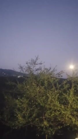 A vertical video pans across a landscape at dusk. The sky is a deep purple, fading to a lighter blue at the horizon. A faint moon is visible in the upper right quadrant of the screen, appearing as a bright, blurry orb. In the foreground, dense green foliage fills the lower half of the frame. To the left of the foliage, a thin, tall, vertical structure, possibly a smokestack or tower, is visible against the sky. Further in the distance, rolling hills or mountains can be seen, and a faint glow of lights suggests a town or city on the horizon. A white guardrail runs along the right side of the frame, with a paved road visible next to it. The overall impression is of a quiet, rural scene transitioning from day to night.