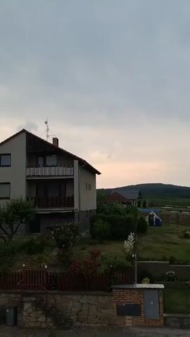 This media file captures a twilight scene in Dobev, Czechia. The focus is on a residential area with several houses and their surrounding gardens. The sky is overcast, indicating either early morning or late evening, with the soft glow of the setting or rising sun faintly visible behind the hills in the distance. The landscape beyond the houses is hilly and appears to be a mix of cultivated fields and natural vegetation.

The foreground features a stone wall with a brick mailbox unit. To the left, a house with a balcony and a chimney stands prominently. To the right, another house with a covered porch and external stairs is visible. The gardens are lush with green grass, bushes, and flowering plants, including roses, adding touches of color to the scene. A small blue and white tent or playhouse is situated in the middle ground of one of the yards. The overall atmosphere is peaceful and quiet, typical of a rural or suburban setting at dusk. No people are visible in the frame. The time of day is suggested by the low light and the muted colors of the sky. The location is identified as Dobev, Czechia, placing the scene in a specific European context.