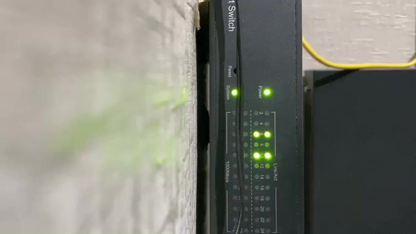 The video shows a close-up of a network switch mounted vertically against a wall. The switch is black and has various LED indicator lights. A green "Power" light is illuminated. Below the power light, there are rows of smaller lights labeled "Link/Act" and numbered ports from 1 to 24. Several of these "Link/Act" lights are also illuminated green, indicating active network connections. A yellow Ethernet cable is visible plugged into the top right side of the switch. The wall beside the switch appears to be textured and light-colored. The focus is on the switch and its indicator lights.