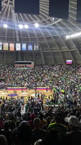 The video shows an aerial view of a crowded basketball arena during what appears to be an intermission or pre-game event. The arena is filled with spectators wearing a lot of green clothing, suggesting a team affiliation. In the center of the court, there are several people on the basketball court, some of whom appear to be holding microphones or performing. There are also several banners hanging from the ceiling of the arena, displaying logos and text, some of which are in Spanish. The court itself has various logos and markings, including a large circular logo in the center. The overall atmosphere appears to be lively and energetic, with a large audience gathered to watch a sporting event.
