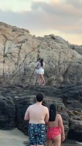 The video captures a marriage proposal taking place on a rocky beach. In the foreground, a shirtless man in floral swim trunks and a woman in a pink one-piece swimsuit are observing the scene. On a rocky outcrop, a couple dressed in white are embracing, seemingly having just gotten engaged. Several other people are visible on the rocks, taking photos or observing the couple. The background features a large rock formation and a sky with a pastel sunset.