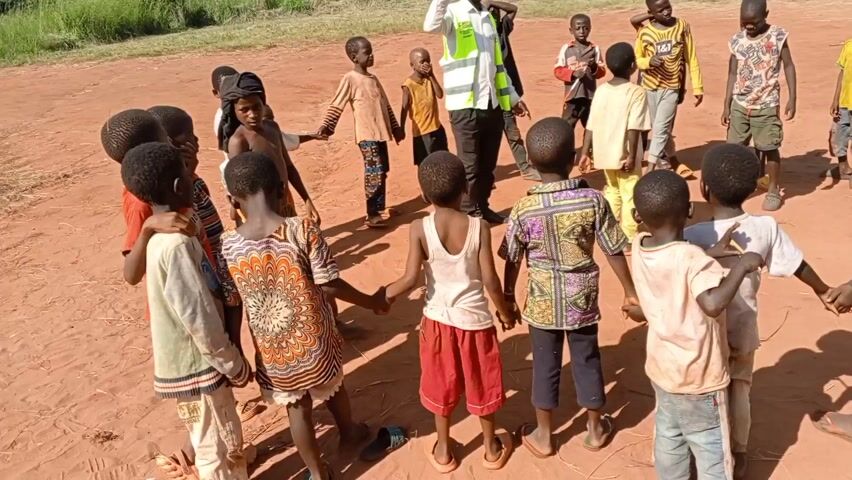 The video shows a group of children gathered on a reddish-brown dirt surface. An adult wearing a yellow safety vest stands among them, gesturing and speaking to the children. The children are of varying heights and dressed in casual clothing, some with bright patterns. One child is holding a small, red object in their hand. Behind them, another child holds up a white banner with text and logos written on it. The background reveals a lush, green landscape with distant mountains. The children seem to be listening to the adult and observing the banner.