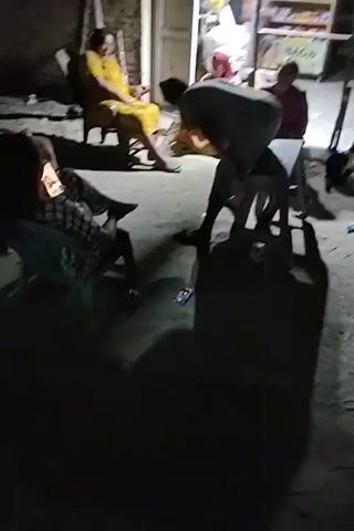 At 00:00, several people are seated outside at night, some on chairs and some on a bench. One person in a yellow dress is holding a phone. At 00:01, the camera pans to the right, showing two individuals sitting on chairs, one in a black t-shirt and the other in a checkered outfit. At 00:02, the camera moves again, focusing on the person in the yellow dress who appears to be talking on the phone. At 00:03, the camera shifts to the right, capturing a person in a gray t-shirt sitting on a bench, with their legs extended. At 00:04, the camera continues to pan right, showing the person in the yellow dress and another individual wearing a pink headscarf and an orange top. The environment appears to be an outdoor setting at night, with dim lighting and some structures in the background.