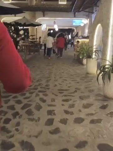 The video shows an outdoor walkway paved with irregular stones, lined with potted plants and what appears to be an outdoor dining area with tables and umbrellas on the left. Several people are walking away from the camera down the walkway. The person closest to the camera, wearing a maroon sweater and blue jeans, is also walking away. The lighting suggests it is evening or a dimly lit area. In the background, there are buildings with white architectural elements and lighting. There is a sign visible in the distance.