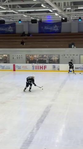 The video shows an indoor ice hockey rink. The ice is smooth and well-maintained. Several hockey players are on the ice, wearing black and white uniforms. Some players are skating with sticks, while others appear to be practicing. In the background, there are bleachers with spectators. There is also a large scoreboard visible, displaying team names and a timer. The overall environment appears to be a professional or semi-professional hockey facility.