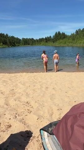 Two children, a girl and a boy, are standing at the edge of a lake on a sandy beach. The girl is wearing a colorful swimsuit, and the boy is wearing swim trunks and a hat. They are facing the water, which is calm and reflects the blue sky. Trees line the opposite shore of the lake, and the sky is a clear, bright blue with a few white clouds. The sun appears to be high in the sky, suggesting it is midday.

A yellow ball is thrown into the water, and both children run into the lake to retrieve it. They splash and play in the shallow water, kicking up water as they move. The scene conveys a sense of carefree fun and summer enjoyment. A dark red blanket or bag is visible in the foreground, partially obscuring the sand. The overall impression is one of a warm, sunny day spent at a tranquil lake.