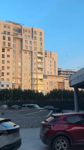 The video shows a tall, modern apartment building under a blue sky. Sunlight reflects strongly off some of the building's windows. The building is a light neutral color, with balconies visible. A line of green evergreen trees is in front of the building, acting as a barrier between the building and a parking area. Several cars are parked in the foreground, including a dark SUV, a dark sedan, and a red hatchback. There is a concrete barrier in front of the cars. The shot is stationary.