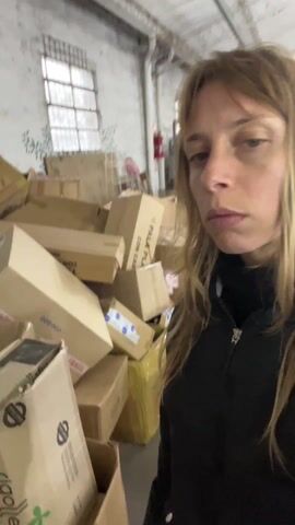 A woman with long, light brown hair and bangs is shown in an indoor setting, likely a warehouse or storage room, in Saladillo, Argentina. She is wearing a black zippered jacket. The space is filled with a large number of cardboard boxes of various sizes, some stacked and others piled. Many boxes feature "FRAGIL" tape. Other visible text on the boxes includes "CON CAN" (partially visible), "TBM," and "020." A window with multiple panes is visible in the background, and a faint green stencil of leaves adorns the wall. The woman looks around the cluttered space, appearing serious or contemplative as she observes her surroundings. The lighting suggests it is daytime.