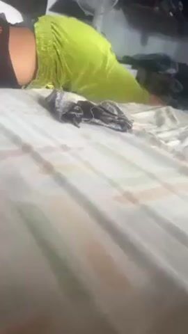 A person wearing a black top and bright green, loose-fitting pants lies face-down on a bed. A small pile of black and silver patterned material rests on the bedspread in front of the person. In the background, a white fan stands on a surface, and a shelf is visible with various items on it. The bed has a patterned white, green, and brown striped bedspread.