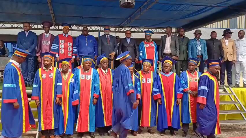 A group of men stands on a stage. Some wear academic gowns in blue, red, and gold, along with academic caps. Others wear suits or casual attire. They are arranged in two rows. A man in a white shirt walks to the front of the group and appears to be interacting with them. The stage is covered with a yellow cloth and set under a blue canopy. A building is visible in the background.