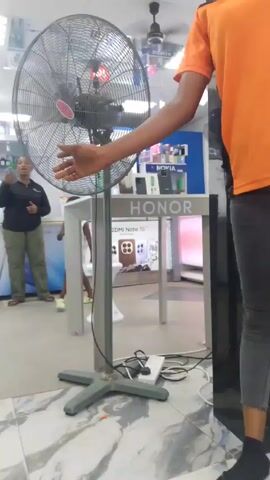 The video shows the inside of a mobile phone store. There is a large standing fan in the foreground with its blades spinning. The camera appears to be inside the store and focused on the fan. A young child in a white dress is seen kicking at the base of the fan. A woman, presumably the child's mother, is standing nearby, holding a phone. Other people, including store employees wearing orange shirts, are visible in the background, near tables displaying phones and accessories. The store has bright overhead lighting and a tiled floor.