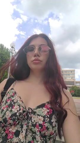 A young woman with long, dark, wavy hair and sunglasses is standing outside, facing the camera. She is wearing a floral print dress with a black background and colorful flowers. Her right hand is raised, and her fingers are visible in the upper left corner of the frame. Behind her, there are trees, a building, and a cloudy sky. The woman is looking directly at the camera and appears to be talking. The scene looks like a casual outdoor setting, possibly a park or a public square.