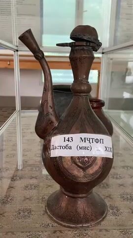 The video shows a close-up of an antique copper ewer with a long, curved spout. The ewer is decorated with intricate engravings. A label attached to the front of the ewer reads "143 MЧТФП Дастоба (мис) а. XIX". The ewer is displayed in a glass case, likely in a museum or exhibition. In the background, other artifacts can be seen, including pottery. The lighting is soft and diffused, creating a warm atmosphere. The camera slowly pans around the ewer, highlighting its details and craftsmanship. The ewer's base is broad and ornate, and the spout curves elegantly upwards. The lid of the ewer is small and metallic, with a decorative element on top. The overall impression is one of historical significance and artistic value.