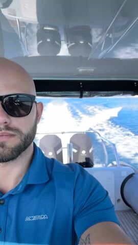 A man with a shaved head and sunglasses is piloting a speedboat. He is wearing a blue collared shirt with "MONTURA" printed on it. Behind him, the boat's wake is visible on the bright blue water. Two boat engines are visible at the back of the boat. The camera is positioned in front of the man, capturing a close-up shot of his face and the surrounding environment.