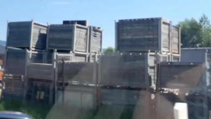 The video displays a series of snapshots of an outdoor industrial or construction site. The first frame shows large, stacked metal cages. The subsequent frames reveal piles of materials, possibly concrete or building components, with a fence in the foreground. A tall, slender smokestack is visible in the background in later frames. The sky is clear and blue. There are no people visible in any of the frames. The overall impression is of a storage or processing area for building materials.