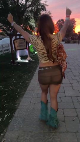 A woman with long brown hair is seen from behind, walking toward the camera, her arms raised. She turns to her right and bends over, looking at the camera. She is wearing a tiger-striped shirt, a short brown skirt, and green boots with tassels. She has a colorful wristband on her left wrist. A brown bag is visible on her back. The background includes a sunset, trees, a sign that reads "Gentle Monster," and other people walking in the distance. There is a brick path and grass on either side of the path. There are inflatable cartoon figures visible in the background.