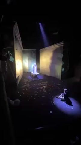The scene is a theatrical production with dark lighting. The stage features cardboard-like structures on either side with the suit of spades on them.  The stage is littered with confetti-like debris.  In the center, a draped figure stands on a small table.  A single beam of light illuminates the figure.  A second figure is seated on the floor with a spotlight on them. The audio consists of male singing.
