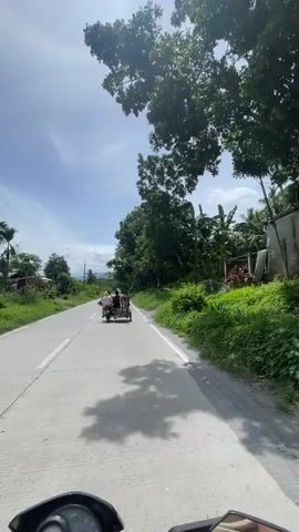 This media file shows a POV shot from a motorcycle traveling on a paved road in Tupi, Philippines. The scene is rural and characterized by lush greenery and trees lining the road. The weather appears to be sunny with scattered clouds, suggesting it is daytime, likely mid-morning or afternoon given the light and shadows.

The primary activity captured is the motorcycle ride itself, following a tricycle ahead. The tricycle is carrying several people, though their specific identities or actions are not clearly discernible. The surrounding environment is dominated by tropical vegetation, including palm trees and dense foliage, indicative of the Philippines' climate. There are also utility poles visible along the side of the road, suggesting the presence of infrastructure.

The road is a two-lane asphalt surface with a white dividing line. The motorcycle's handlebars and a portion of its front are visible at the bottom of the frame, reinforcing the first-person perspective. The overall mood conveyed is one of a leisurely journey through a scenic, natural landscape. No visible text is present in the media file.