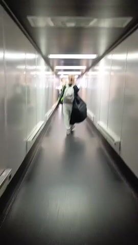 The media file shows two people walking down a jet bridge. The person in front is wearing a white outfit, a light gray backpack, and carrying a large black bag over their shoulder. They have blonde hair. Behind them, a second person is visible, also carrying a backpack. The jet bridge has white, reflective walls and a dark floor. Bright fluorescent lights run along the ceiling, illuminating the corridor. The overall lighting suggests it is daytime, though no windows are visible to confirm the time of day or weather conditions. The scene appears to be in an airport, likely on the way to or from an airplane. The people are walking away from the camera, so their expressions are not visible, but the context of travel implies a sense of movement and transition. No text is visible in the media file. The location context provided is Saburovka, Russia, which could indicate this jet bridge is at an airport serving that region.