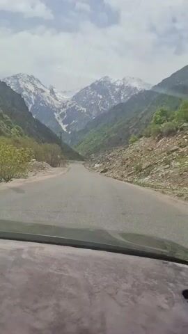 A car drives down a paved road through a mountainous valley. The mountains are covered in green trees on the lower slopes, and patches of snow at higher elevations. The road is clear and in good condition. The sky is partially cloudy, with some patches of blue. Power lines run along the road. The interior of the car is visible at the bottom of the frame. Music is playing.