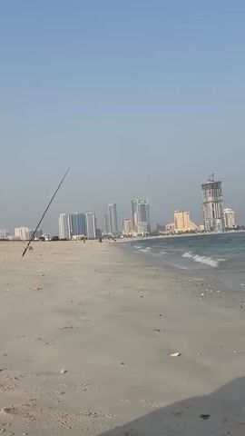 The video is a vertical, first-person shot of a sandy beach with buildings in the background. The ocean is visible on the right, with small waves lapping at the shore. A fishing rod is planted in the sand on the left side of the frame. The sky is clear and blue. The buildings appear to be modern high-rises, some of which are under construction. The overall impression is a serene coastal scene with urban development.