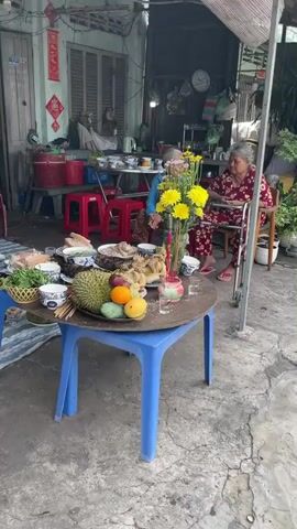 The video shows an outdoor scene in front of a house in Vietnam. A blue plastic table sits on a concrete floor and is covered in various food offerings including a whole durian, mango, grilled fish, boiled chicken, and bowls of soup. A vase of yellow flowers and a small pot of burning incense sit on the table. Two elderly women are seated in the background. One woman is sitting in a chair with a walker next to her wearing a red floral outfit. The other woman is sitting next to her wearing a blue jacket and patterned dress. She smiles at the camera. Red plastic stools are scattered around the scene. The house is visible in the background.