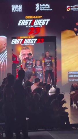 The video shows a stage at the “East West Armwrestling 23” event in Germany. The crowd is silhouetted as they watch three men enter the stage, walking from behind a large screen. The screen displays the event logo and a portrait of a man. The three men on stage are wearing matching “East West” jerseys.