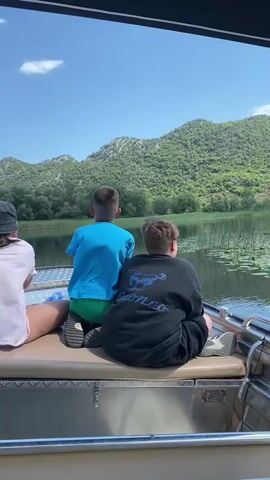 The video shows three individuals from the back, seated on a boat. The person on the left wears a white shirt and a black hat. The middle person is wearing a blue shirt and green shorts, and the person on the right is wearing a black sweatshirt. They are looking out at a body of water with green hills and trees in the background. The sky is blue with some clouds. The boat appears to be moving.