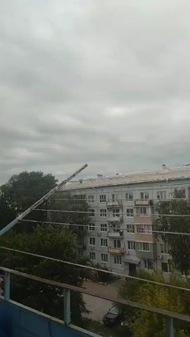 A video shows a cloudy sky over a residential building. A long pole-like object extends from the left side of the frame, angled upwards towards the sky, and passes in front of the building. The building is light gray with multiple floors and windows, some with balconies. Trees are visible in front of and to the side of the building. The overall lighting suggests an overcast day.
