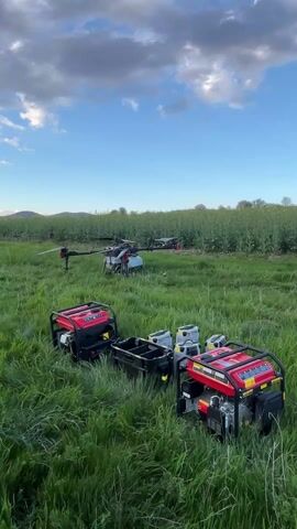 A drone is taking off in a grassy field. The drone is a quadcopter type, colored gray, with a mounted liquid tank underneath. Behind the drone stand two red generators and a black container holding several battery packs. In the background is a field of green plants and a blue sky with white clouds.
