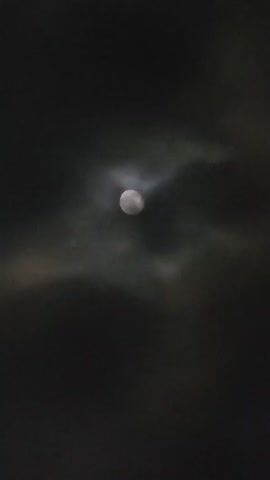 The video shows a close-up of the moon partially obscured by clouds. The moon is bright and circular, with a reddish-orange hue. Wispy clouds drift across the moon, creating a soft, ethereal effect. The background is dark and black, suggesting it is nighttime. The video appears to be filmed at an angle, giving the impression of the moon moving through the sky.