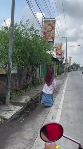 A person wearing a maroon hijab and a light-colored patterned top with a long blue skirt is walking away from the camera along the side of a paved road. The person is on a concrete walkway next to a ditch. Several poles with banners are visible along the road. The banners are red and yellow with text and images that are difficult to discern but appear to be advertisements. Trees and buildings line the street. The camera appears to be mounted on a motorcycle, as the handlebars and a red mirror are visible in the foreground. The sky is overcast.