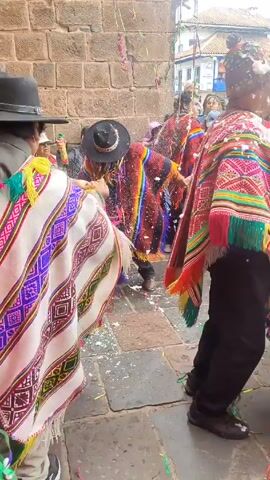 A street celebration unfolds in a town, possibly in South America, evidenced by the town's architecture and the traditional clothing worn by the participants. The scene is festive, with colorful balloons strung overhead and confetti littering the ground. Many people are dancing. The participants are wearing vibrant, traditional clothing, including woven shawls and hats. Some people hold hands and dance in pairs, while others observe the celebration. The setting appears to be a town square, with buildings visible in the background. There are many spectators watching the dance.
