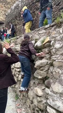 A group of young people is climbing on and around an old stone wall. Some are at the top of the wall, others are ascending, and some are at the bottom waiting their turn. The wall is constructed of rough, gray stones and has some vegetation growing out of it. In the background, a stone tower is visible, suggesting the wall is part of a historical structure. The weather appears overcast. The individuals are dressed casually in various types of clothing, including jackets, jeans, and sneakers. They seem to be enjoying themselves and helping each other.