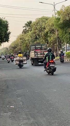 The video shows a street scene with several motorbikes and a truck. The motorbikes are all traveling in the same direction on a paved road lined with trees. Many of the motorbikes have passengers. One motorbike in the foreground has a person wearing a green jacket and black pants, and another person behind them. Another motorbike has a passenger with a light brown headscarf. A large green truck is visible on the right side of the screen. The sky is overcast.