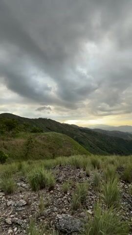 The video captures a panoramic view of a mountainous landscape under a cloudy sky. The foreground is dominated by rough, rocky terrain sparsely covered with dry grass and small green shoots. In the middle ground, rolling hills covered in lush green vegetation stretch into the distance. The background reveals a valley with scattered settlements and more distant, hazy mountains. The sky is filled with dramatic, dark gray clouds, with some lighter patches suggesting the presence of sunlight filtering through. The camera is stationary, offering a static view of the scene. There are no people or visible actions within the frame.