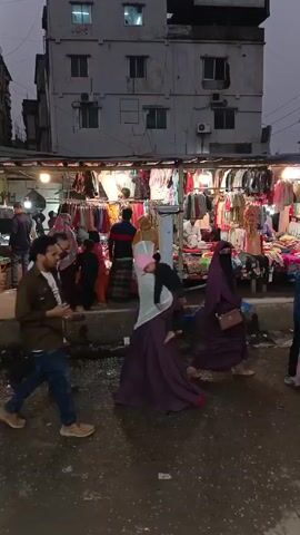 The video shows a busy street market in what appears to be an urban area in Asia. Several people are walking around and browsing through clothing displayed for sale under a row of lighted stalls. An auto rickshaw, carrying several female passengers, drives through the scene, honking its horn. The street appears somewhat muddy. Tall buildings are visible in the background. Most people appear to be pedestrians.