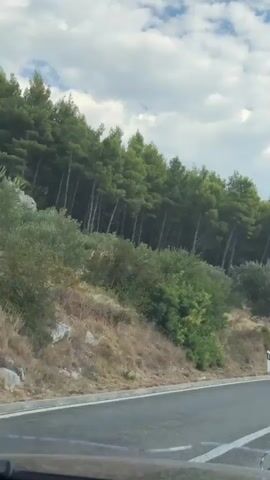 The video shows a road with a steep embankment covered in trees and vegetation on the right side. The road is paved and appears to be a highway or a main road, with lane markings visible. In the distance, a gray SUV is driving on the road towards the camera. The sky is blue with scattered white clouds. The overall scene suggests a scenic drive through a natural landscape.