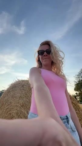 A woman with blonde hair and sunglasses, wearing a pink tank top and denim shorts, is in a field with hay bales. She is holding a camera out in front of her, as if taking a selfie. The sun is setting in the background, casting a warm glow over the scene. The sky is blue with some clouds. The field appears to be a harvested wheat field.