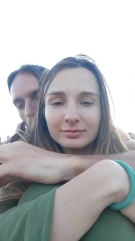 A low-angle, close-up video shows a couple embracing outdoors, with a bright sky in the background. A man with long brown hair is behind a woman with long brown hair, his arms wrapped around her. They are both wearing green clothing. The man is holding a wooden flute to the woman's mouth as if she is playing it. The woman has her eyes closed and a slight smile. In the background, slightly out of focus, there are other people in festive clothing. The lighting suggests it is daytime, possibly late afternoon or early evening.