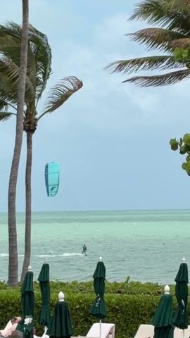 Here's a detailed description of the video:

The scene is set at a beach resort with a view of the ocean. Palm trees are visible on either side, framing the shot. In the foreground, there's a row of closed green beach umbrellas and lounge chairs, suggesting a poolside or beachfront area.

In the distance, a person is kitesurfing. The kite is turquoise blue with a white logo, clearly visible against the slightly overcast sky. The ocean water is a muted green color. The waves are gentle, and the kitesurfer appears to be moving across the water at a moderate speed. 

The sky is predominantly light blue with some cloud cover, creating a diffused light. The overall atmosphere is relaxed and summery, despite the lack of bright sunshine.