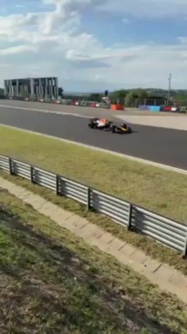 A race car drives around a track, followed by the camera panning to a group of people standing beside a race track, observing a pink and white car. The people are dressed casually. There are white tents in the background.