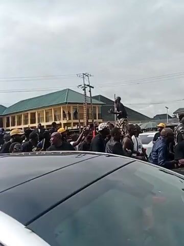 The video shows a funeral procession with a large crowd of people dressed in black. A red vehicle is visible with a casket on top, draped in white fabric and adorned with flowers. Some individuals are standing on the vehicle. People are walking alongside the vehicle, and some are riding on motorcycles. The environment appears to be an outdoor setting with buildings in the background and an overcast sky. The overall mood seems somber.