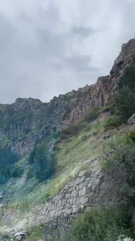 This is a video of a person or people in a vehicle traveling along a mountain road. The road is partially constructed with stone walls on the side. The mountainside is covered in green vegetation and rocks. The sky is cloudy. The camera is looking out from inside the vehicle, possibly through a wet window, which causes some blurring and streaks in the footage. The vehicle appears to be moving, as indicated by the shifting perspective of the landscape.
