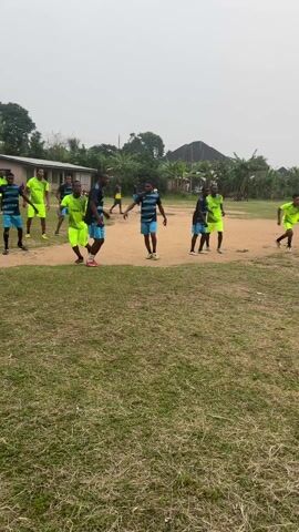 A soccer team wearing blue and black striped jerseys with turquoise accents and another team wearing neon yellow and black striped jerseys are playing a soccer game on a dirt field. Several players jump near the goal to try to score a goal with a white soccer ball. A person standing near the camera watches the action, while buildings and lush trees are visible in the background. The sky is overcast.